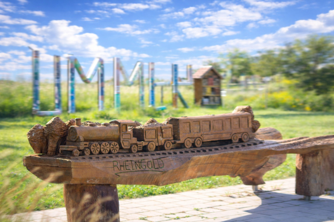 Die hölzerne Sitzbank „Rheingold“ am Rheinvorland in Germersheim zeigt einen historischen Zug und verbindet Kunsthandwerk mit regionaler Verkehrs- und Industriegeschichte.